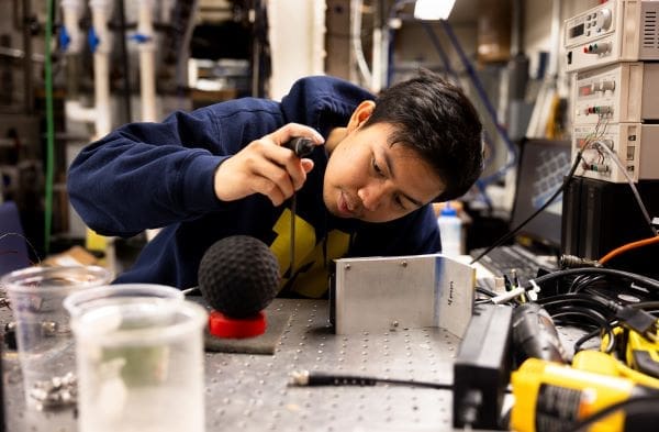 Image: A man sits at a steel workbench with a soft-ball sized sphere resting on a platform placed in front of him. He holds a t-shaped tool a few inches away from the sphere and peers at it from the side (s. smart vehicle)