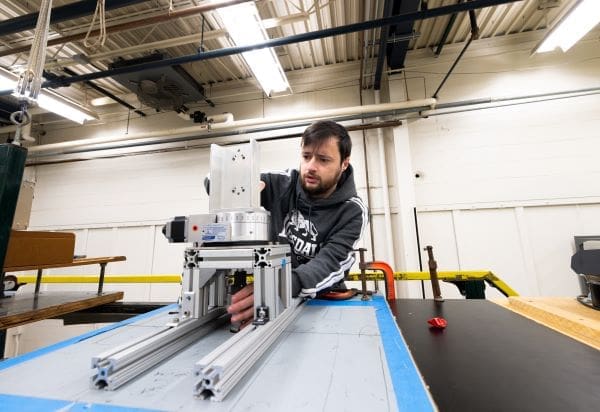 Image: A man stands behind a black bench with his gaze focused on his right hand as it adjusts a silver device, positioned on top of sliding rails (s. smart vehicle)