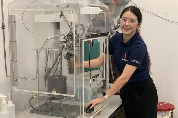 Image: A woman in a blue t-shirt and lab glasses is standing in a laboratory demonstrating a process