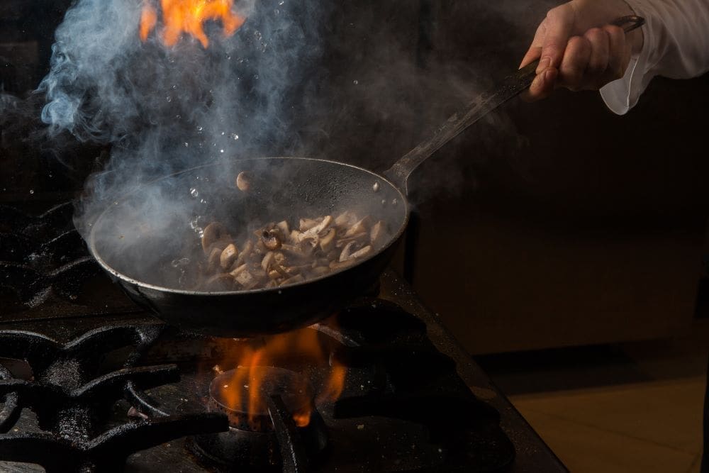 Image: Side view mushroom frying with smoke and fire and human hand and pan in stove