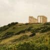 Image: large stone structure sitting on top of a lush green hillside -Sounion, Greece