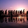Image: A line of silhouetted people walking along a waterfront at sunset with reflections on the water