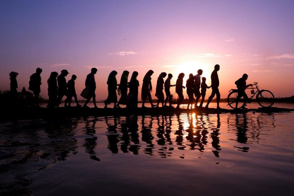 Image: A line of silhouetted people walking along a waterfront at sunset with reflections on the water